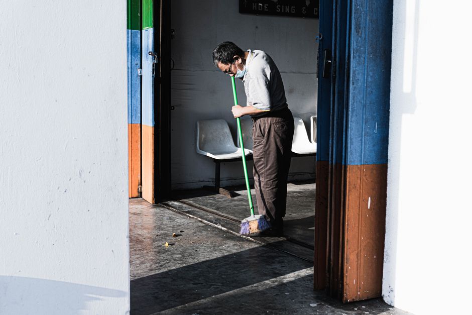 A person performing surface cleaning on a dark carpeted floor in a dimly lit room, using a long-handled cleaning tool, with a large white projection screen overhead and black partition walls in the ba