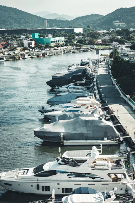 A scenic view of a calm lake surrounded by lush green hills under a partly cloudy sky, with three boats docked at a floating yellow and blue pontoon dock system. The boats include a white speedboat wi