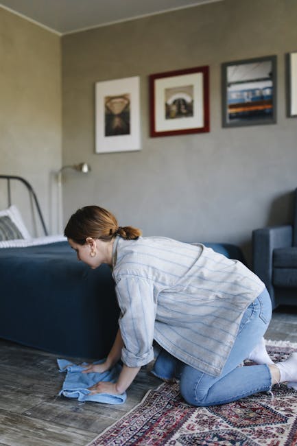 A woman with brown hair tied back, dressed in a light striped shirt and jeans, kneeling on a patterned rug in a living room while using a blue cloth to clean the dark wooden floor. The room features a