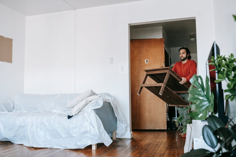 A woman seated on a white couch covered with a protective sheet in a residential living room, with a man kneeling at the base of the couch, holding the sheet to prepare for cleaning. The room features